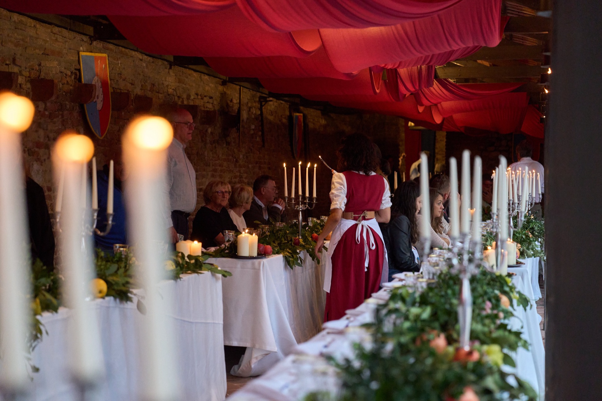 Banchetto rinascimentale alla Taverna del Barbacane: lunghe tavolate con tovaglie bianche, candelabri accesi, drappi rossi al soffitto e damigella in costume del Casalino